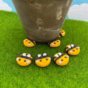 May include: A selection of yellow bee-shaped decorations with black and white stripes and pink cheeks. The bees are arranged on green artificial grass, with a brown pot in the background.