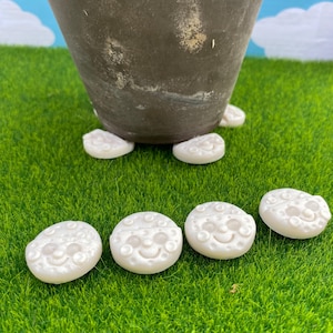 May include: White, moon-shaped decorative stones with smiling faces are scattered on green artificial grass. A dark brown pot sits atop the stones. The stones have a pearl-like finish and are arranged around the base of the pot.