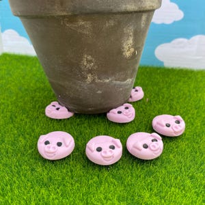 May include: A brown ceramic flower pot with pink pig-shaped pot feet. Several additional pink pig feet are scattered on the green surface. The pig feet have black eyes and smiling faces.