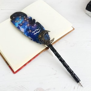 May include: A decorative pen with a feather-shaped top featuring a celestial design in shades of blue, purple, and white. The pen has a black, textured handle and a metal tip. The pen is resting on an open notebook.