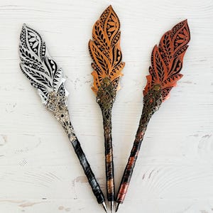 Könnte beinhalten: Drei dekorative Federstifte mit aufwendigen Designs. Einer ist silber und weiß, ein anderer orange und schwarz, und der dritte rot und schwarz. Jeder Stift hat ein detailliertes Metallakzent und einen gemusterten Griff.