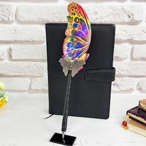 May include: A black pen featuring a large, vibrant, multi-colored butterfly wing topper with a metallic bronze-toned butterfly accent at the base of the wing. The pen is held upright by a small, rectangular black base stand.