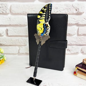 May include: A black pen featuring a large, detailed butterfly wing topper in yellow, black, and blue, accented with a metallic bronze butterfly charm at the base of the wing. The pen stands upright on a small, rectangular black base.