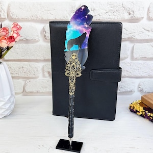 May include: A decorative pen with a feather-shaped top featuring a wolf silhouette against a galaxy design. The pen has a black base and a detailed gold filigree accent. A black notebook and a vase of pink roses are in the background.