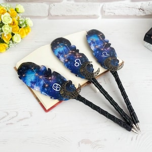 May include: Three pens featuring dark blue galaxy-patterned feathers with gold constellation designs and white zodiac symbols for Cancer, Leo, and Libra. Each pen has a black wrapped handle and a decorative bronze-coloured crescent moon accent.