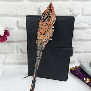 May include: A decorative feather pen featuring detailed patterns in orange, brown, and black, presented on a black stand. It's positioned in front of a black notebook. A vase of flowers is to the left. The pen's design is intricate.