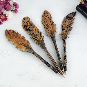 Puede incluir: Cuatro bolígrafos decorativos con forma de pluma y diseños intrincados. Las plumas son doradas y negras, con patrones detallados. Los mangos de los bolígrafos son negros y dorados, con adornos metálicos ornamentados. Los bolígrafos están dispuestos sobre una superficie de madera blanca.
