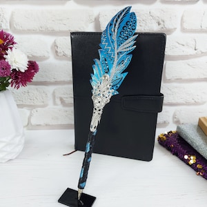 Puede incluir: Un bol&iacute;grafo de pluma ornamental con un dise&ntilde;o azul y plateado. El bol&iacute;grafo presenta una parte superior decorativa en forma de pluma con detalles intrincados y una base de metal plateado. El bol&iacute;grafo se exhibe sobre un soporte negro.