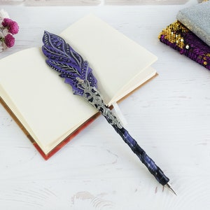 Peut inclure: Un stylo décoratif doté d'un embout en forme de plume violette et noire avec des détails en filigrane argenté. Le manche est enveloppé d'un ruban violet foncé et noir, menant à une pointe en argent métallique. Le stylo repose sur un carnet ouvert.