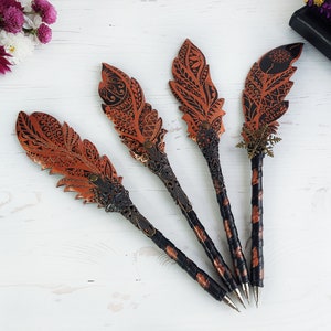 May include: Four pens featuring decorative feather-shaped tops with intricate black and copper-colored patterns. Each pen has a black wrapped handle and a metallic tip. One pen includes a small bronze-colored snowflake charm near the feather.