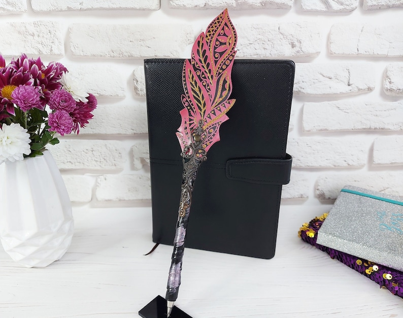 Puede incluir: Un bol&iacute;grafo decorativo con una parte superior en forma de pluma rosa y dorada. El bol&iacute;grafo tiene un cuerpo a rayas negras y plateadas y se apoya en un soporte negro. Un cuaderno negro y un jarr&oacute;n de flores est&aacute;n en el fondo.