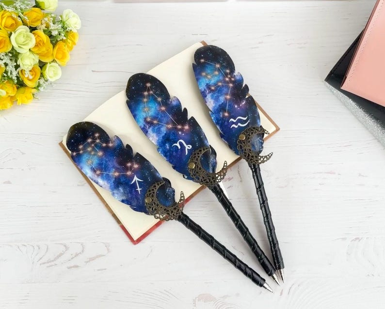 May include: Three feather-shaped pens with celestial designs. The pens feature a blue and purple galaxy pattern with constellations and zodiac symbols. The pen handles are black with a textured design, and the pen tips are silver.