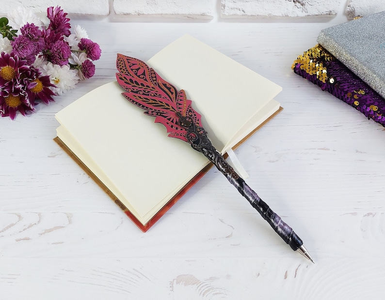 Puede incluir: Un bol&iacute;grafo decorativo con una parte superior en forma de pluma. La pluma es rosa con dise&ntilde;os negros grabados. El cuerpo del bol&iacute;grafo est&aacute; envuelto en tela negra y morada. El bol&iacute;grafo descansa sobre un cuaderno abierto con p&aacute;ginas en blanco.