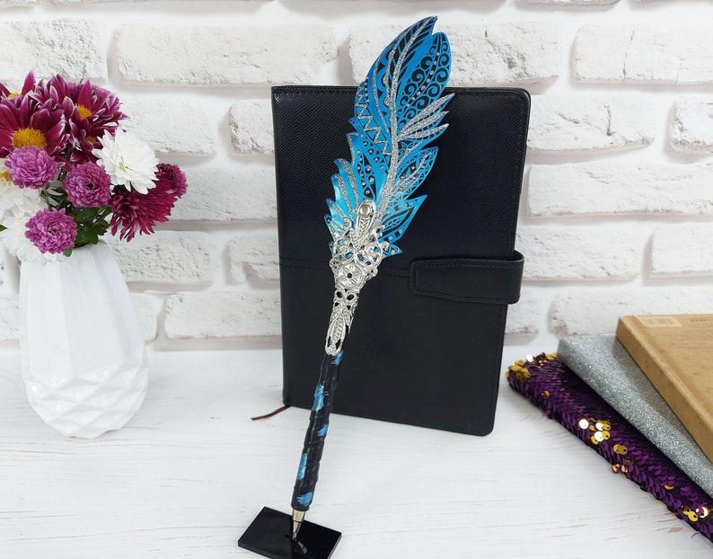 Puede incluir: Un bol&iacute;grafo de pluma ornamental con un dise&ntilde;o azul y plateado. El bol&iacute;grafo presenta una parte superior decorativa en forma de pluma con detalles intrincados y una base de metal plateado. El bol&iacute;grafo se exhibe sobre un soporte negro.