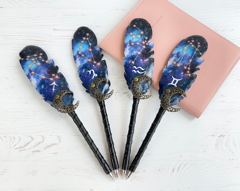 May include: Four decorative pens with feather-shaped tops featuring a celestial design. Each pen has a different zodiac constellation and symbol, with a bronze-colored crescent moon accent. The pen handles are black and the background includes a pink notebook.