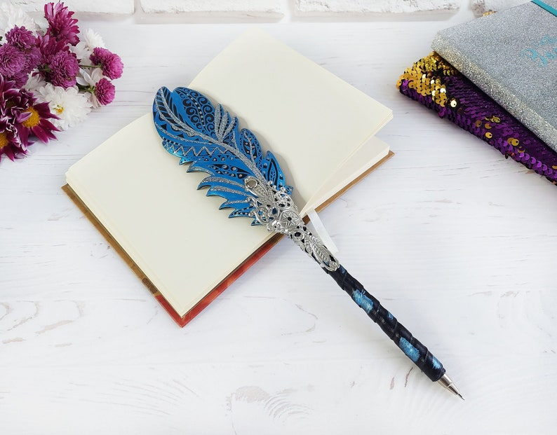 Puede incluir: Un bol&iacute;grafo decorativo con forma de pluma azul y plateada descansa sobre un cuaderno abierto. El bol&iacute;grafo presenta una parte superior detallada en forma de pluma con patrones intrincados y una base de metal plateado. El mango del bol&iacute;grafo es negro con detalles en azul. Un ramo de flores est&aacute; en el fondo.