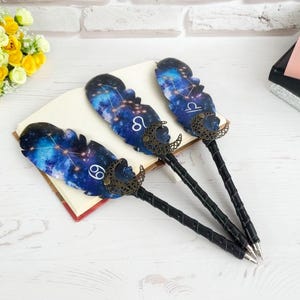 May include: Three decorative pens with feather-shaped tops featuring a galaxy design and zodiac symbols. The pens have black, textured handles and bronze-colored accents. The pens are resting on a book.
