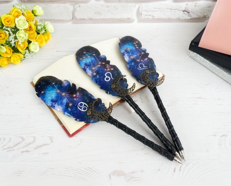 May include: Three decorative pens with feather-shaped tops featuring a galaxy design and zodiac symbols. The pens have black, textured handles and bronze-colored accents. The pens are resting on a book.