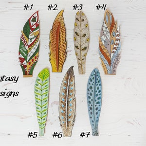 May include: Seven decorative feather designs with various patterns and colours. The feathers include green, red, yellow, brown, and blue hues. Some feathers have the text "Happy Holidays". The words "Fantasy Designs" are also visible in the image.