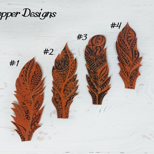 Puede incluir: Cuatro recortes decorativos de plumas de varios tama&ntilde;os, cada uno con intrincados patrones negros sobre un fondo naranja quemado. Las plumas est&aacute;n dispuestas sobre una superficie de madera blanca. El texto "Copper Designs" est&aacute; en la esquina superior izquierda.