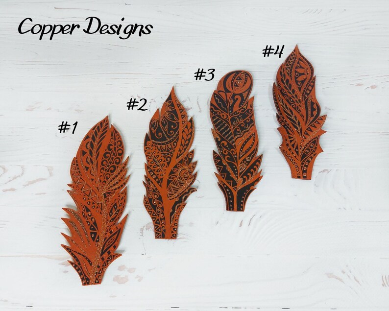 Puede incluir: Cuatro recortes decorativos de plumas de varios tama&ntilde;os, cada uno con intrincados patrones negros sobre un fondo naranja quemado. Las plumas est&aacute;n dispuestas sobre una superficie de madera blanca. El texto "Copper Designs" est&aacute; en la esquina superior izquierda.