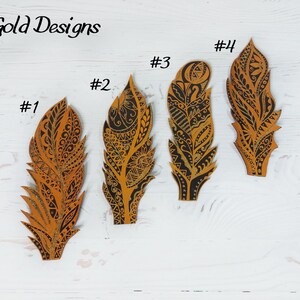 Puede incluir: Cuatro adornos decorativos en forma de pluma con intrincados dise&ntilde;os negros y dorados. Las plumas est&aacute;n numeradas del 1 al 4, y el texto "Gold Designs" est&aacute; en la parte superior. Las plumas est&aacute;n sobre una superficie de madera blanca.