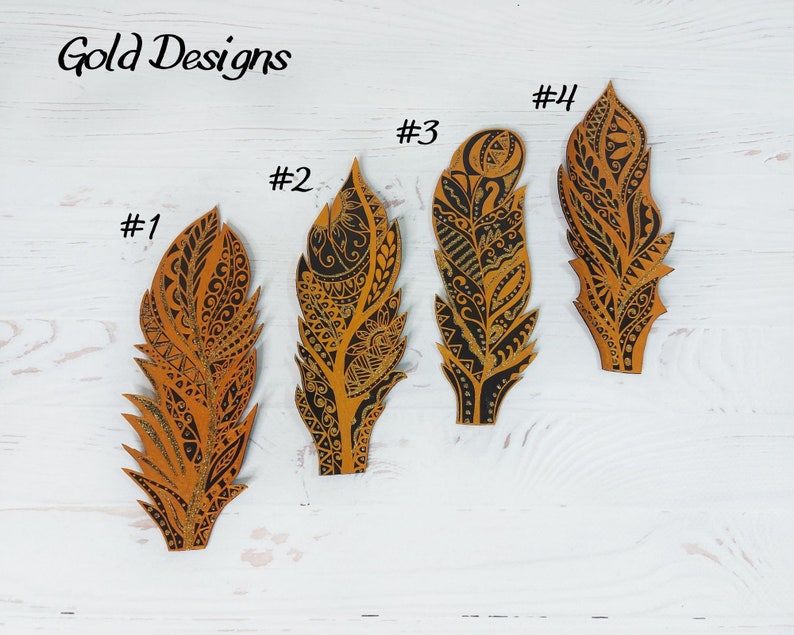 Puede incluir: Cuatro adornos decorativos en forma de pluma con intrincados dise&ntilde;os negros y dorados. Las plumas est&aacute;n numeradas del 1 al 4, y el texto "Gold Designs" est&aacute; en la parte superior. Las plumas est&aacute;n sobre una superficie de madera blanca.