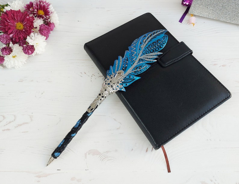 Puede incluir: Un bol&iacute;grafo decorativo con dise&ntilde;o de pluma azul y detalles plateados descansa sobre un diario de cuero negro. El bol&iacute;grafo presenta detalles intrincados y un agarre texturizado. Un ramo de flores blancas y moradas est&aacute; en el fondo.