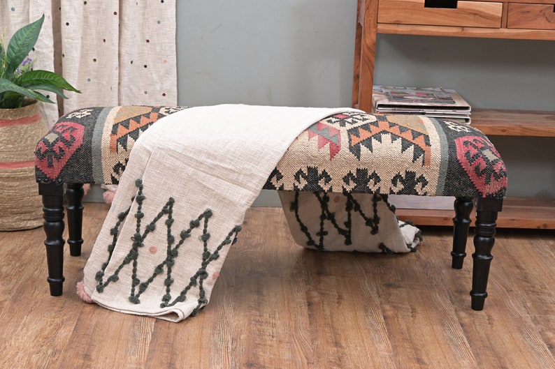 K&ouml;nnte beinhalten: Dekorative Bank mit gemustertem Sitz in Beige-, Schwarz-, Rot- und Oranget&ouml;nen. Die Bank hat schwarze, gedrechselte Beine und ist mit einer cremefarbenen Decke mit dunkelgr&uuml;nen und rosa Akzenten drapiert. Wohnkultur.