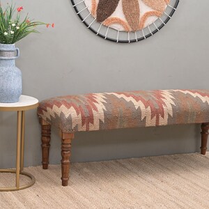 K&ouml;nnte beinhalten: Eine gepolsterte Bank mit einem geometrischen Muster in Rot-, Beige- und Braunt&ouml;nen. Die Bank hat gedrechselte Holzbeine. Ein Beistelltisch mit wei&szlig;er Platte und goldenen Beinen steht neben der Bank. Eine Vase mit Blumen steht auf dem Tisch.