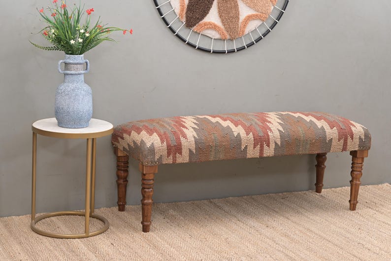 K&ouml;nnte beinhalten: Eine gepolsterte Bank mit einem geometrischen Muster in Rot-, Beige- und Braunt&ouml;nen. Die Bank hat gedrechselte Holzbeine. Ein Beistelltisch mit wei&szlig;er Platte und goldenen Beinen steht neben der Bank. Eine Vase mit Blumen steht auf dem Tisch.