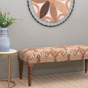 K&ouml;nnte beinhalten: Dekorative Bank mit einem Sitz aus Stoff mit Mustern in Braun-, Gr&uuml;n- und Oranget&ouml;nen. Die Bank hat vier Holzbeine. Eine blaue Vase mit Blumen steht auf einem kleinen Beistelltisch. &Uuml;ber der Bank befindet sich ein runder Wandbehang.