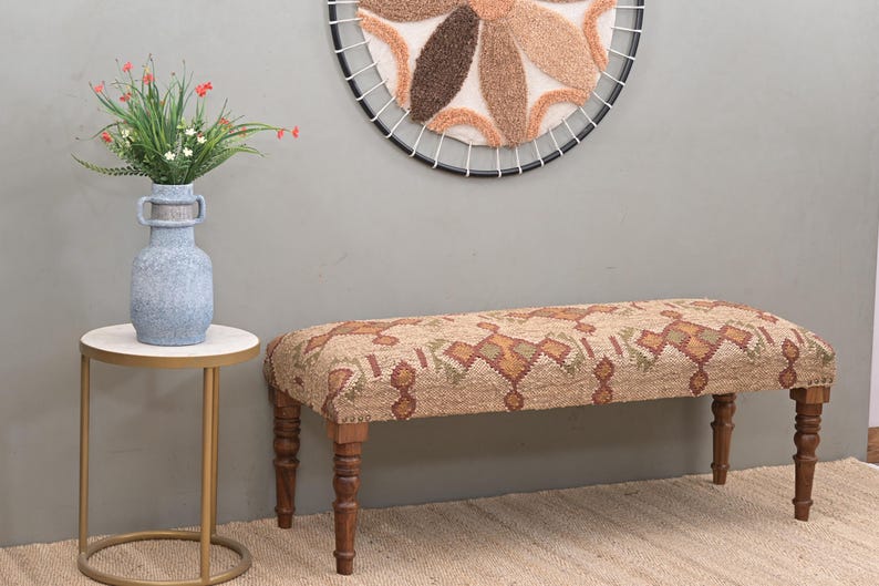 K&ouml;nnte beinhalten: Dekorative Bank mit einem Sitz aus Stoff mit Mustern in Braun-, Gr&uuml;n- und Oranget&ouml;nen. Die Bank hat vier Holzbeine. Eine blaue Vase mit Blumen steht auf einem kleinen Beistelltisch. &Uuml;ber der Bank befindet sich ein runder Wandbehang.