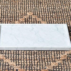 May include: A rectangular white marble tray featuring subtle grey veining throughout the surface. The tray has a smooth, flat base and slightly raised edges, providing a clean and simple design for holding various small household items.