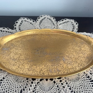 May include: A gold-colored oval serving tray with a floral pattern. The tray has handles on each end and is resting on a white lace doily. The tray's surface is covered in a delicate, repeating floral design.