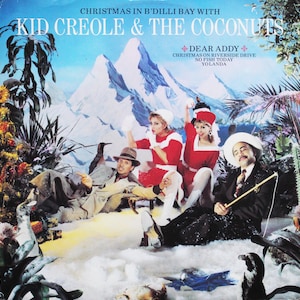 Puede incluir: Una portada de álbum de vinilo vintage de Kid Creole & The Coconuts. La portada representa una escena de montaña nevada con cuatro personas vestidas con ropa de invierno. El título del álbum es "Christmas in B'Dilli Bay with Kid Creole & The Coconuts".
