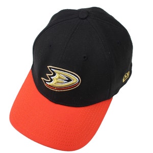 Puede incluir: Una gorra de béisbol negra y naranja con un logotipo bordado en oro de un pájaro estilizado con las alas extendidas. El logotipo tiene un contorno blanco y la gorra tiene una visera negra y una corona naranja.