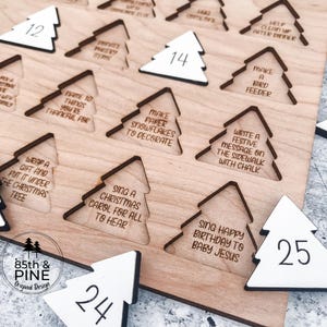 May include: Wooden Christmas advent calendar with tree-shaped cutouts and numbered white tree-shaped pieces. Each cutout has a festive activity. The calendar includes the text "85th & Pine Original Design."