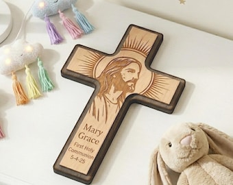 May include: Wooden cross with a detailed engraving of Jesus Christ, surrounded by a sunburst design. The cross has a dark brown border and includes the inscription "Mary Grace, First Holy Communion 5-4-25". A plush bunny and cloud-shaped lights with tassels are also in the image.