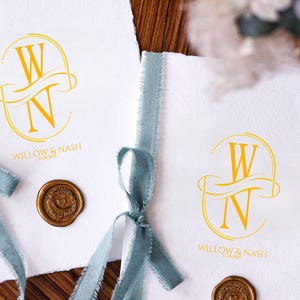 Puede incluir: Dos tarjetas blancas con un monograma dorado de las letras "W" y "N" est&aacute;n atadas con cintas azul claro. Las tarjetas tienen un sello de cera dorado con un dise&ntilde;o de coraz&oacute;n. El texto "WILLOW & NASH" est&aacute; impreso en las tarjetas.