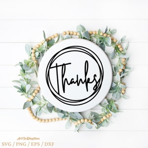 May include: A white round sign with the word "Thanks" written in black cursive lettering inside a double circle. The sign is surrounded by a garland of green leaves and beige beads.