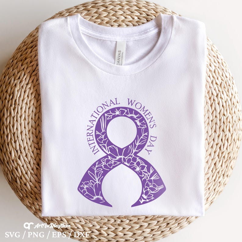 以下が含まれることがあります： 白いTシャツに、花柄のディテールが施されたスタイリッシュな数字の8の紫色のデザインが施されています。数字の8の周りに「国際女性デー」の文字が書かれています。