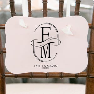 Puede incluir: Un letrero de silla rosa claro con un monograma negro de las letras "F" y "M" dentro de un dise&ntilde;o ovalado. El letrero tambi&eacute;n incluye los nombres "Faith & Mavin" y la fecha "06.28.2015" en texto negro. El letrero est&aacute; sujeto a una silla de madera.