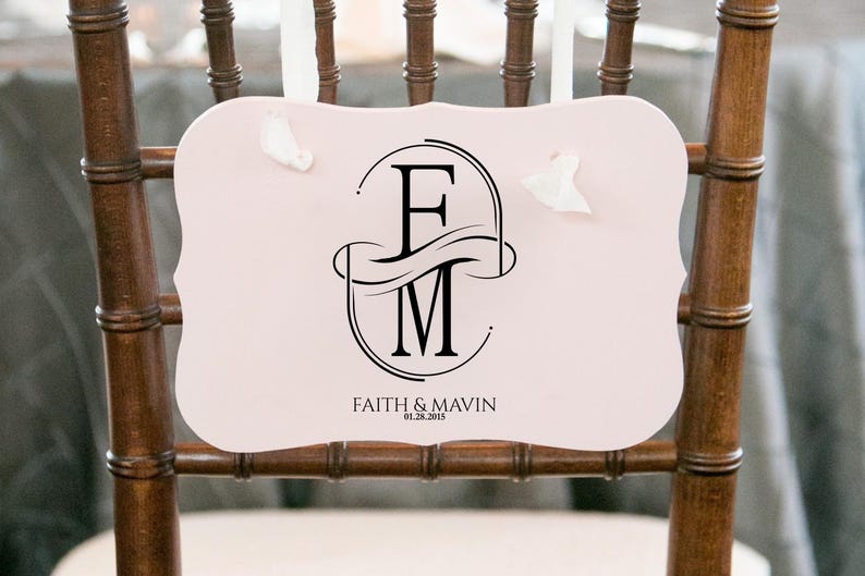 Puede incluir: Un letrero de silla rosa claro con un monograma negro de las letras "F" y "M" dentro de un dise&ntilde;o ovalado. El letrero tambi&eacute;n incluye los nombres "Faith & Mavin" y la fecha "06.28.2015" en texto negro. El letrero est&aacute; sujeto a una silla de madera.