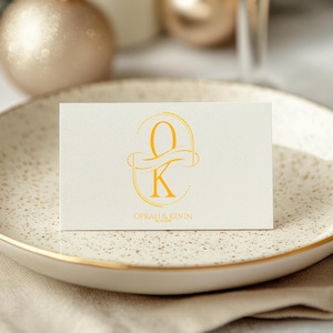 Puede incluir: Una tarjeta color crema con letras doradas y las iniciales "O" y "K" se muestra en un plato moteado con borde dorado. La tarjeta tambi&eacute;n incluye los nombres "Oprah & Kevin". El plato descansa sobre una servilleta de lino beige.