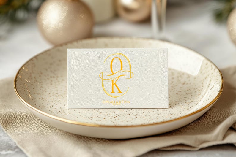 Puede incluir: Una tarjeta color crema con letras doradas y las iniciales "O" y "K" se muestra en un plato moteado con borde dorado. La tarjeta tambi&eacute;n incluye los nombres "Oprah & Kevin". El plato descansa sobre una servilleta de lino beige.