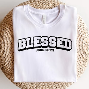 May include: White t-shirt with the word "BLESSED" in a black, distressed font, arched across the chest. Below the word "BLESSED" is the text "JOHN 20:29" in a smaller font. The shirt is displayed on a woven, circular surface.