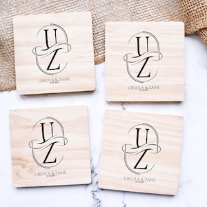 Puede incluir: Cuatro posavasos cuadrados de madera clara. Cada uno presenta un monograma negro con las letras "U" y "Z" entrelazadas, los nombres "URSULA & ZANE" y una fecha. Los posavasos est&aacute;n dispuestos sobre una superficie blanca.