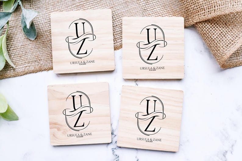 Puede incluir: Cuatro posavasos cuadrados de madera clara. Cada uno presenta un monograma negro con las letras "U" y "Z" entrelazadas, los nombres "URSULA & ZANE" y una fecha. Los posavasos est&aacute;n dispuestos sobre una superficie blanca.