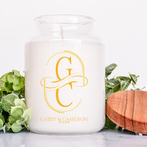 Puede incluir: Una vela blanca en un tarro de cristal con tapa de madera. La vela tiene un monograma dorado y el texto "Gabby & Cameron" con una fecha. El follaje verde est&aacute; en el fondo.